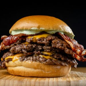 Mouth-watering triple bacon cheeseburger with avocado sauce on a wooden board against a dark background.